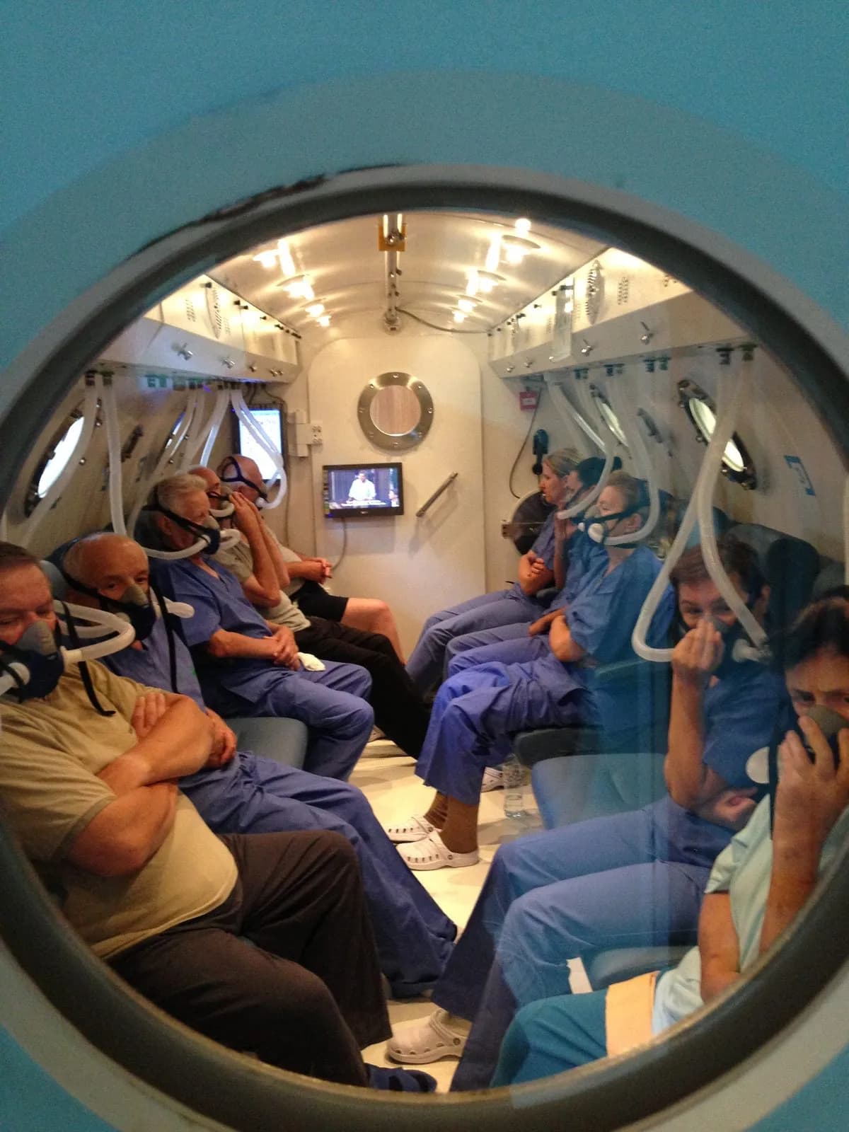 Looking through the round viewport of a multiplace chamber door during a session: two rows of seated occupants in blue scrubs, each wearing a mask on a breathing hose