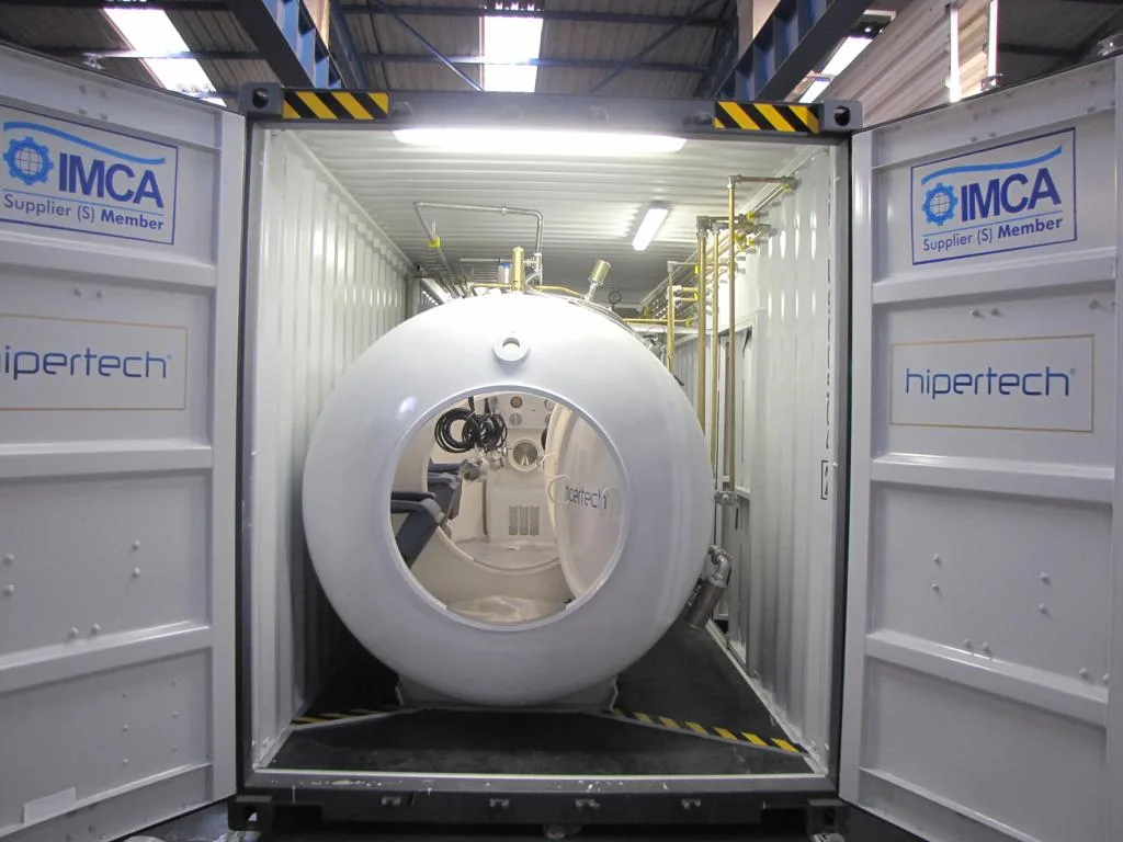 The doors of a Hipertech containerized system standing open, the white chamber inside seen end-on through its open lock door, IMCA Supplier (S) Member decals on both doors