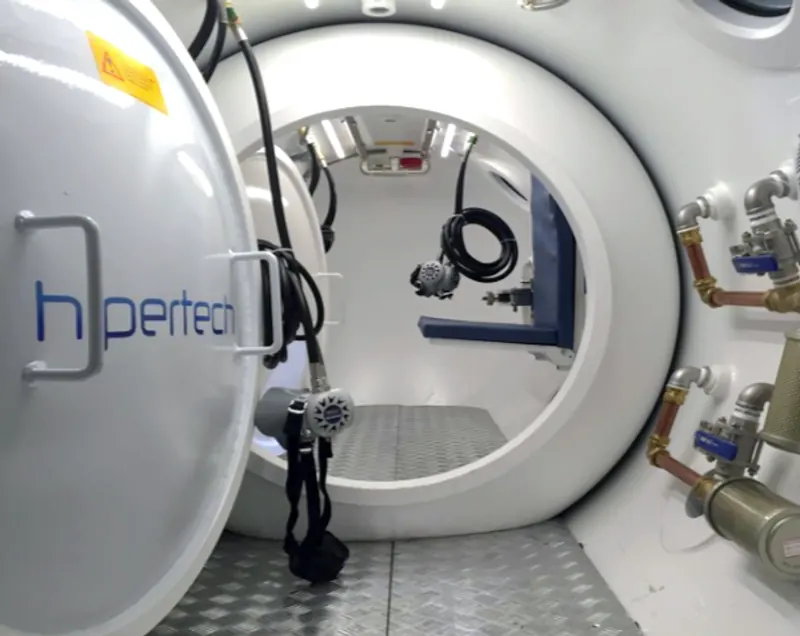 The open door of a chamber inside its container, the Hipertech wordmark on the door leaf and a mask harness hanging within