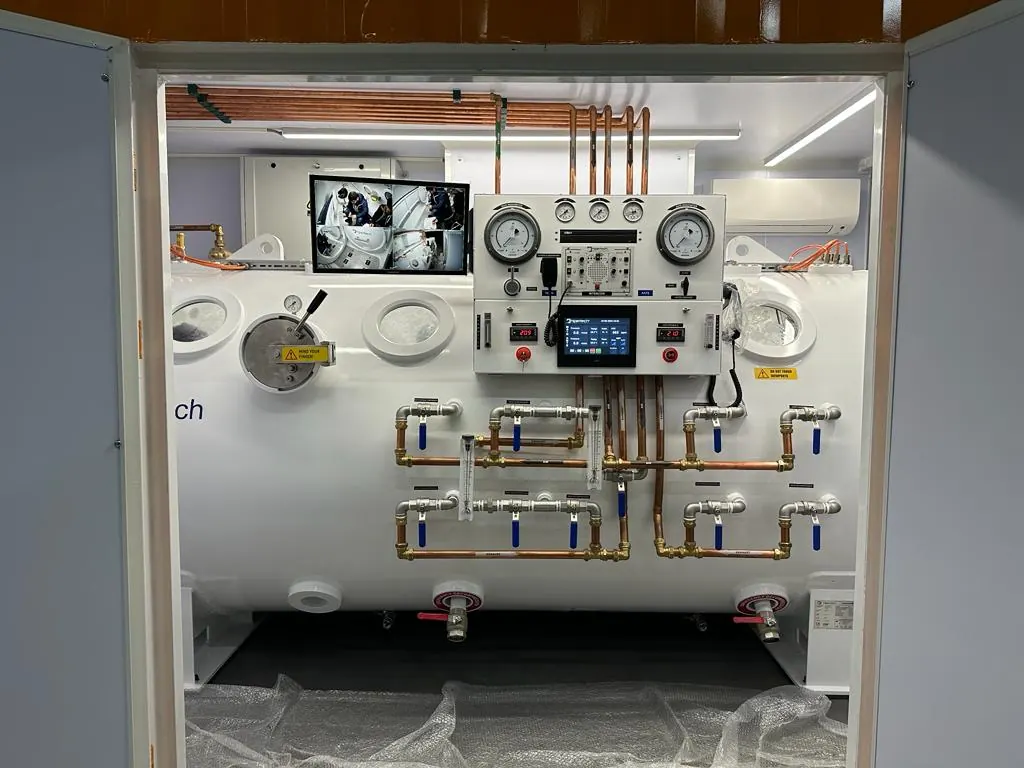 The console face of a containerized chamber seen through the open container doors: two depth gauges, a touchscreen, a monitor and a brass valve manifold