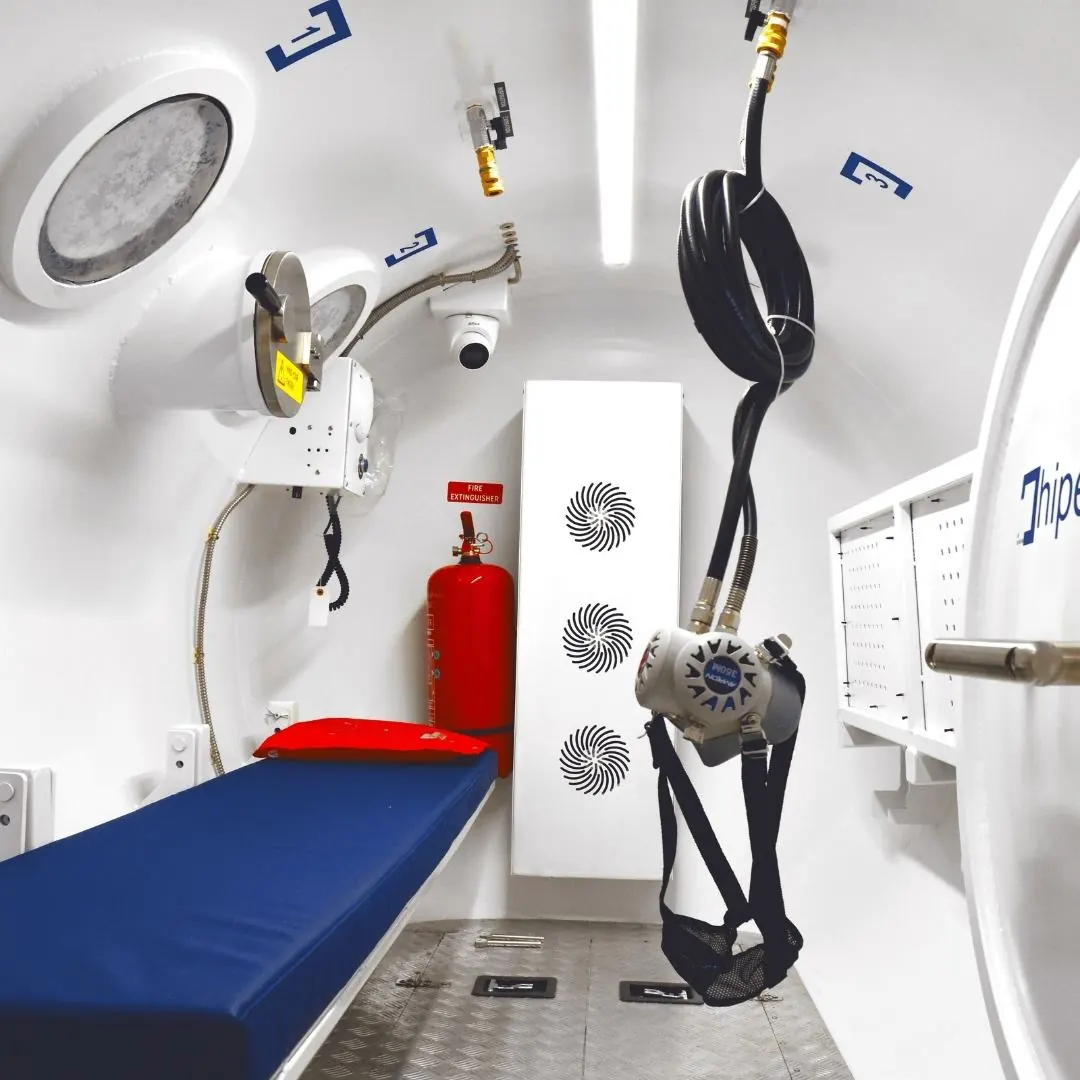 Inside a containerized chamber: a padded bench, a fire extinguisher, a scrubber column with three fan grilles, a mask on its harness and a dome camera