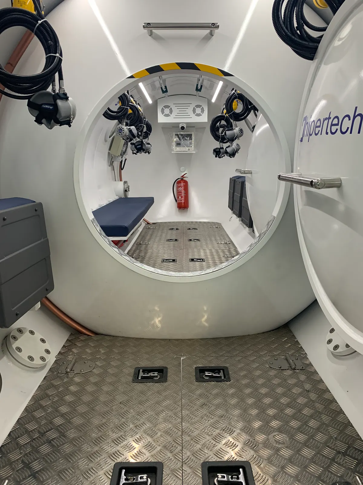 Looking through the open outer door of a decompression chamber into its entry lock: a navy padded bench, with masks and hoses on the bulkhead