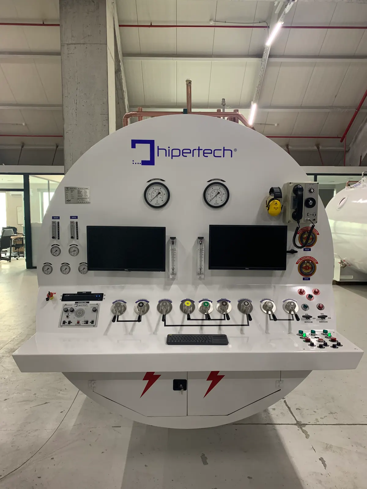 A twin-lock decompression chamber head-on: the circular white end, two depth gauges, two monitors and a wide console shelf of valve levers