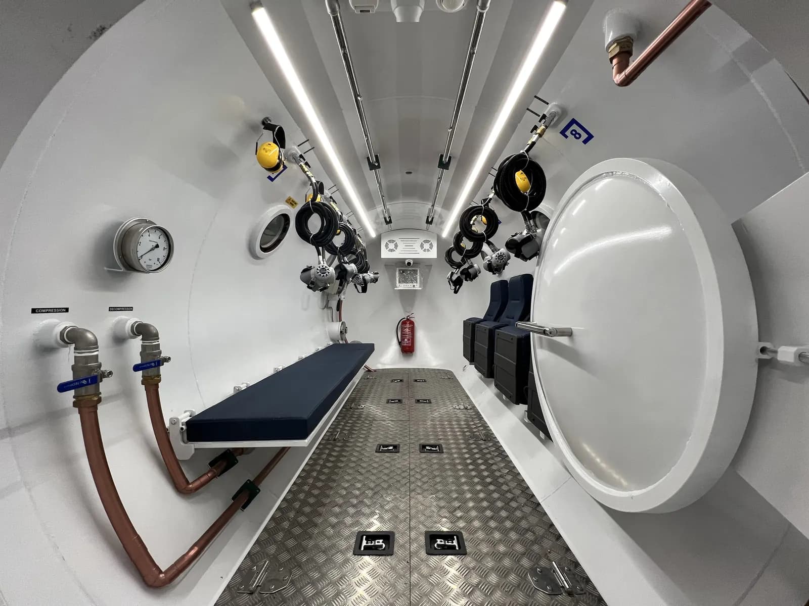 Inside a decompression chamber: a padded bench one side, folding seats the other, masks and coiled hoses on both bulkheads