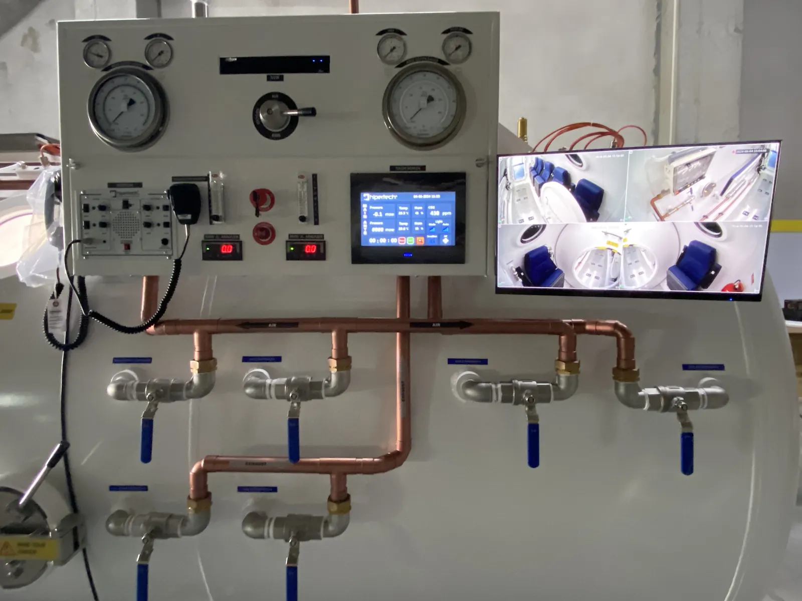 A decompression chamber control panel: depth gauges, a touchscreen, a monitor showing the interior, and the valve manifold running along the hull below