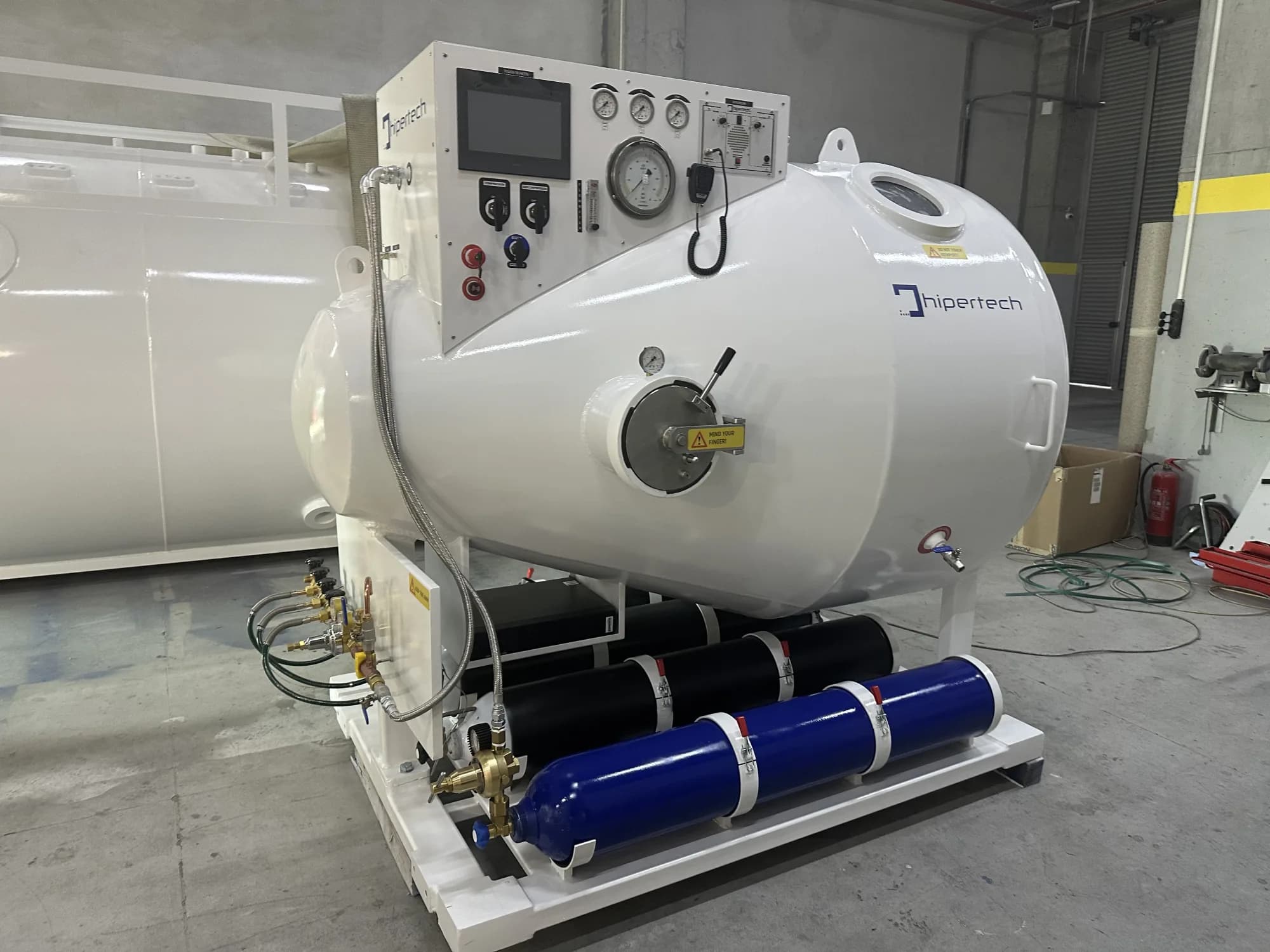 A Hipertech transportable recompression chamber on a white skid, black and blue composite gas cylinders mounted under the frame and a touchscreen control console on the angled face of the hull