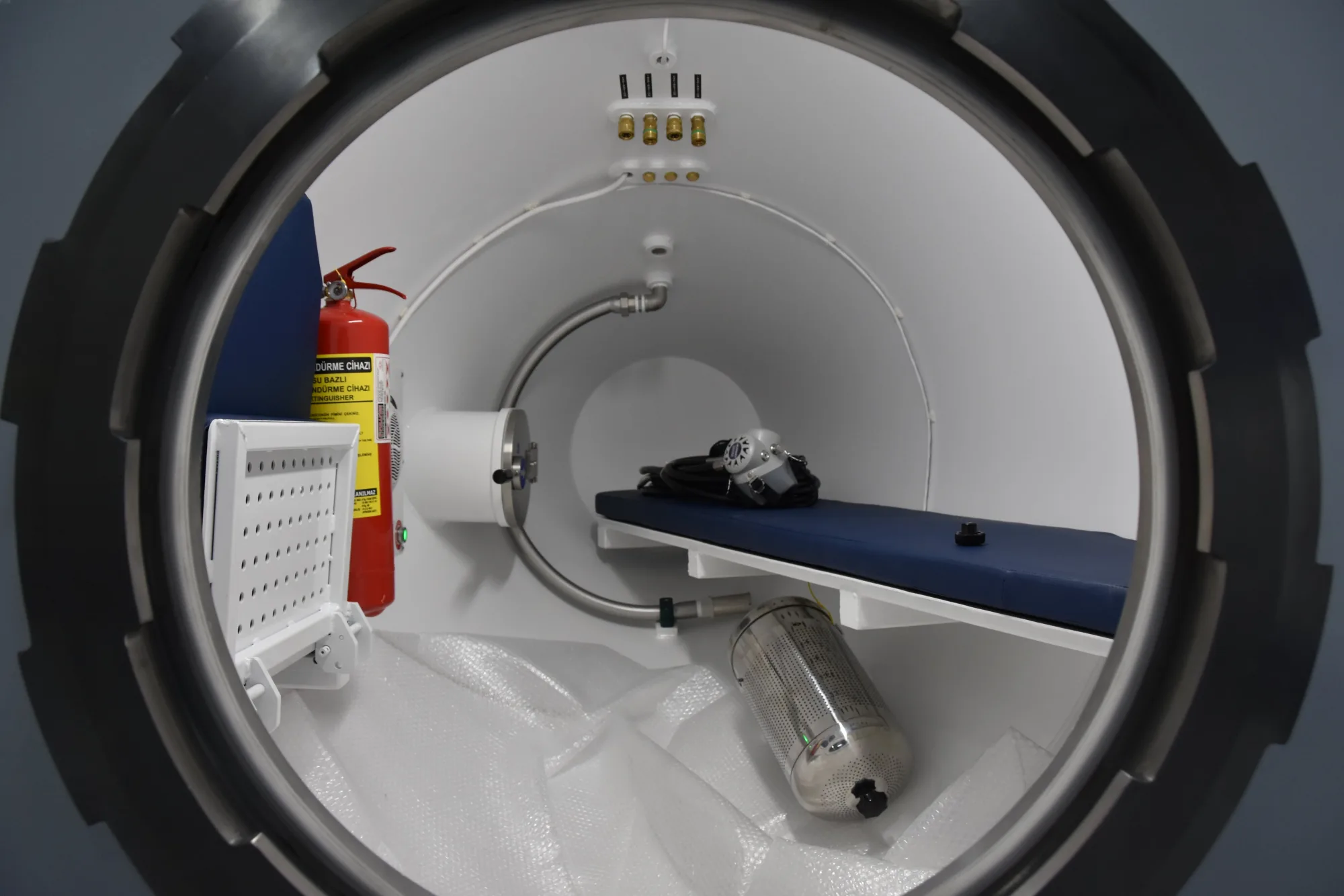 Looking through the open door of a transportable recompression chamber: a padded bench, gas ports and a fire extinguisher inside