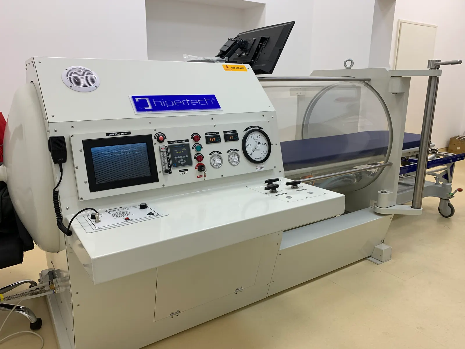 A Hipertech monoplace chamber in a clinical room: a clear acrylic hull, a blue mattress inside, and a control head carrying gauges and a touchscreen