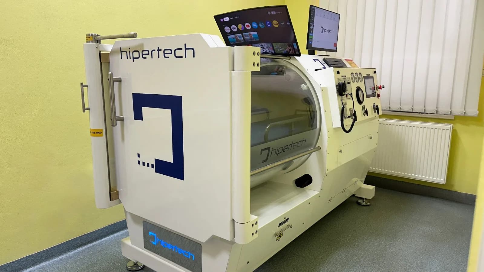 A Hipertech monoplace chamber installed in a hospital room: a cream shell with its door swung open, the clear acrylic hull behind it, and a control head with gauges and two screens