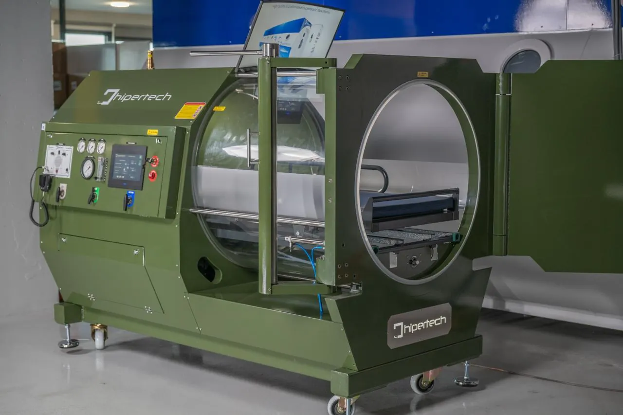 An olive-green monoplace chamber with its door open and its console alongside, on the factory floor