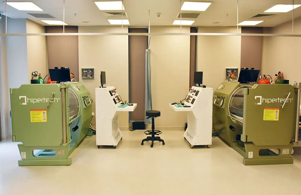 Two olive-green monoplace chambers facing each other across a clinical room, each with its own control trolley