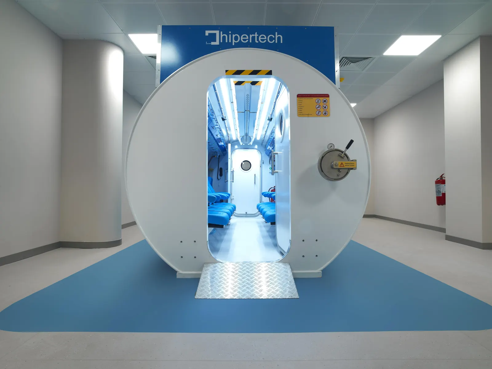 A Multiplace Omega seen end-on down a hospital corridor, its oval door standing open on a lit interior of blue seats, a checker-plate ramp to the sill