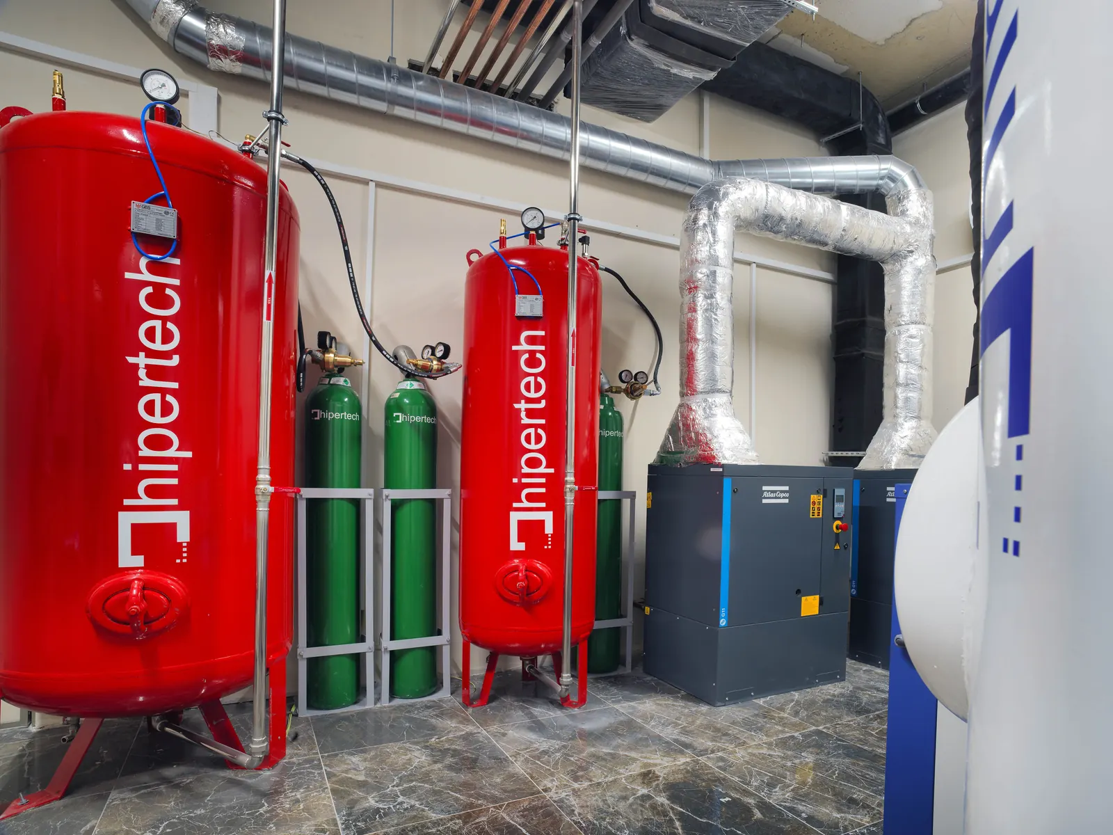 A hyperbaric centre plant room: two red Hipertech pressure vessels, a rack of green gas cylinders and lagged ductwork, with the chamber hull at the edge of frame