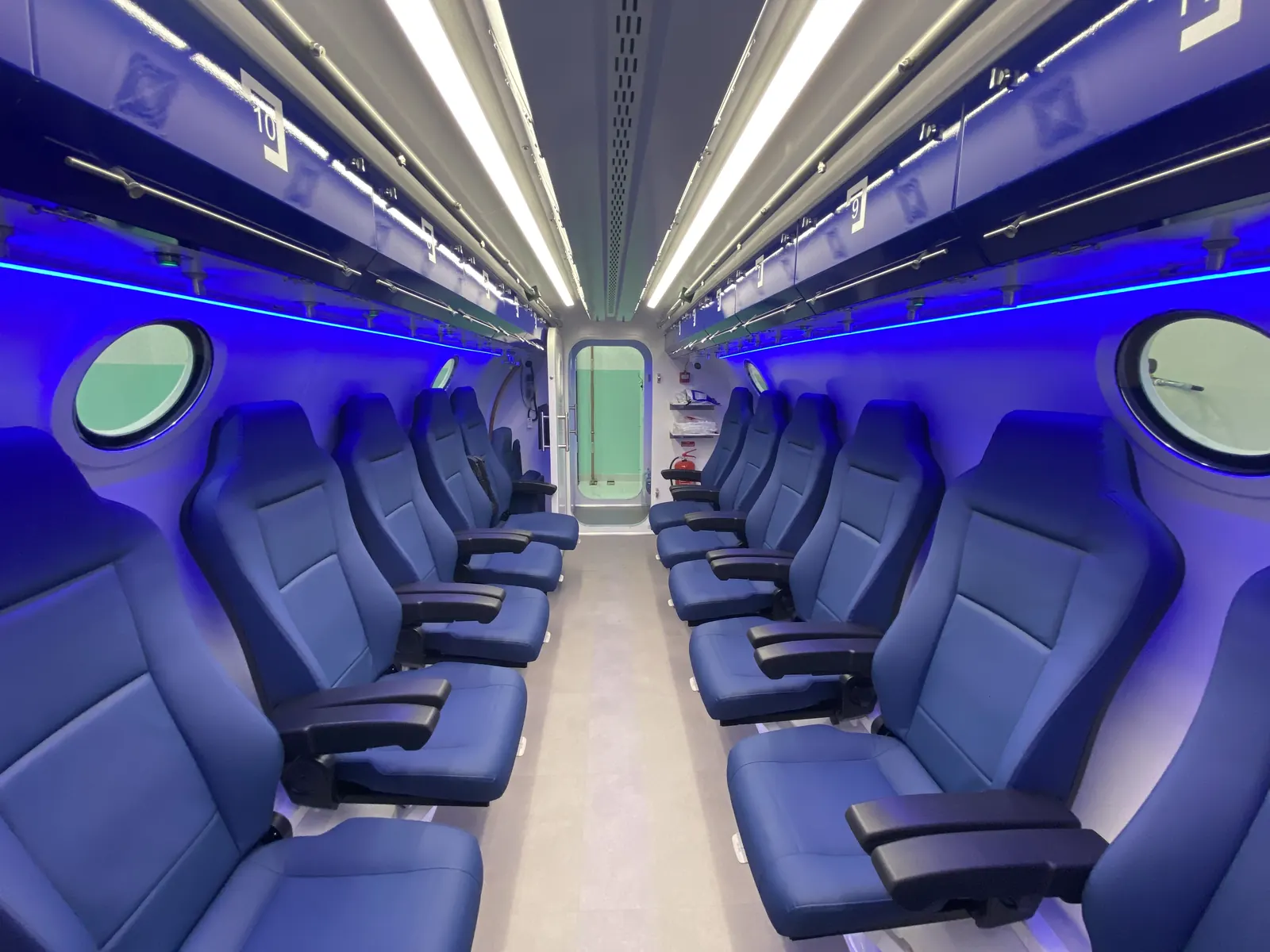 A Multiplace Omega interior under blue cove lighting, two rows of seats and green-tinted oval viewports either side