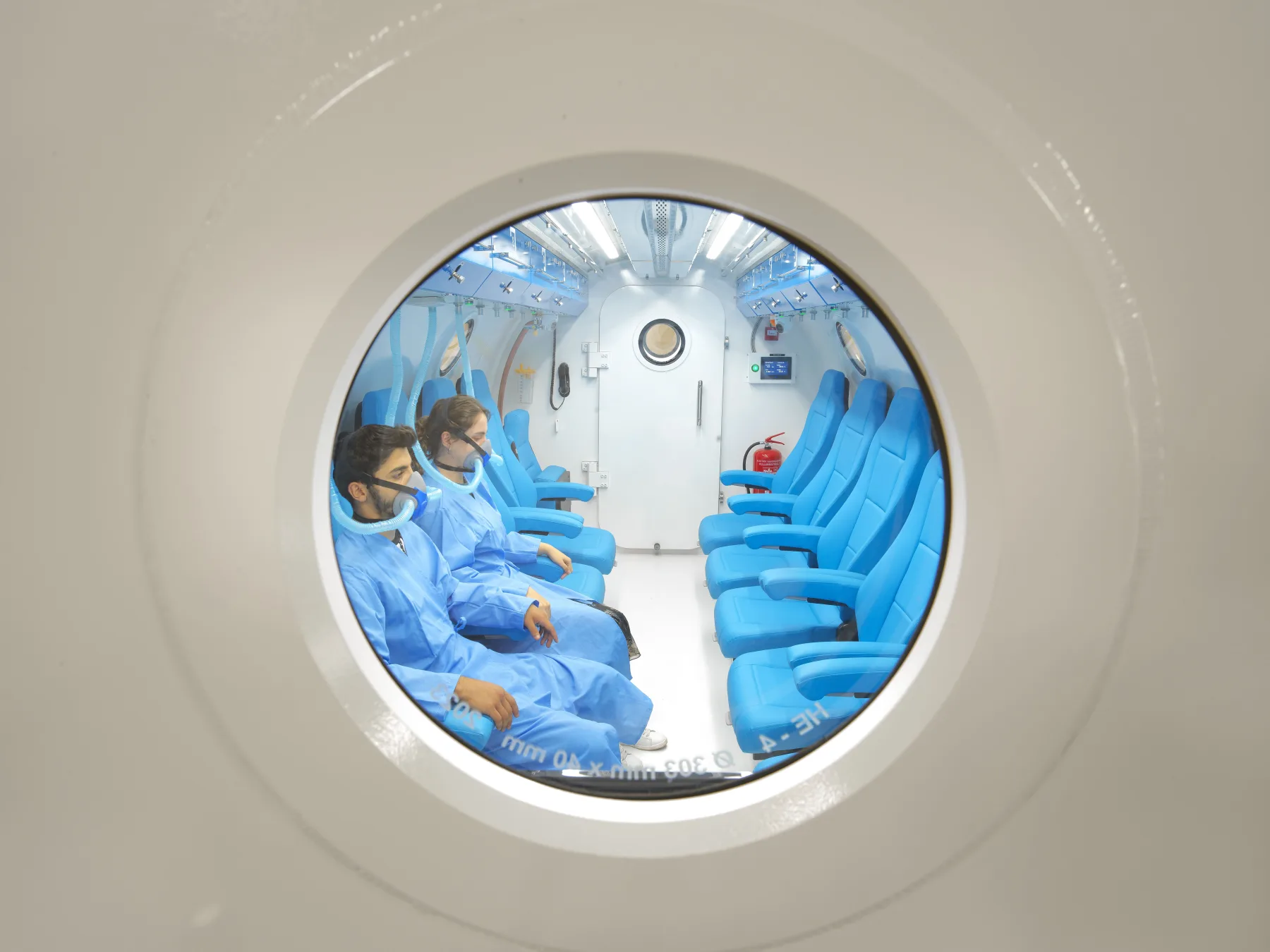 Looking through the open round door of a Multiplace Omega: two rows of blue seats down a lit aisle, with two people in blue scrubs seated inside