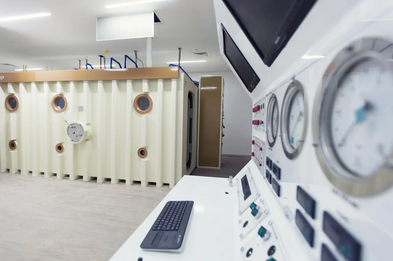A Multiplace Quadro seen across its control room from the operator's console: depth gauges and a keyboard in the foreground, the chamber's cream fluted cladding, bronze-rimmed viewports and door beyond