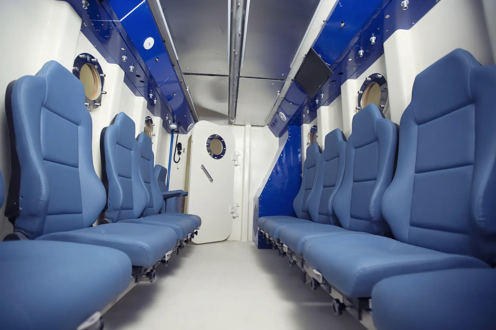 Inside a Multiplace Quadro: pale blue seats down both sides with a fold-down bench, round side viewports and a rectangular inner lock door
