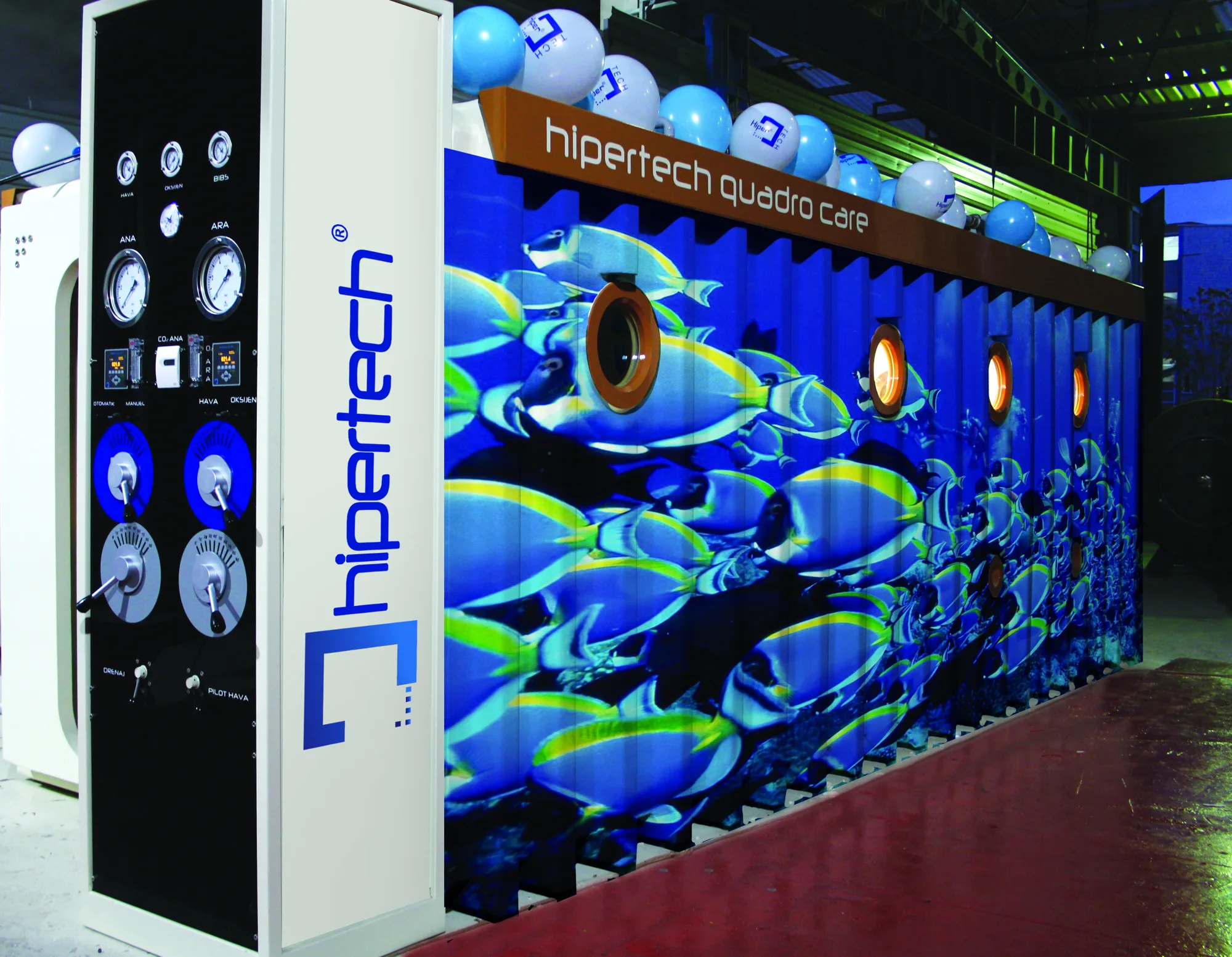 A Multiplace Quadro with its white control console in front, beside the chamber wrapped in a tropical fish mural under a lit hipertech quadro care header