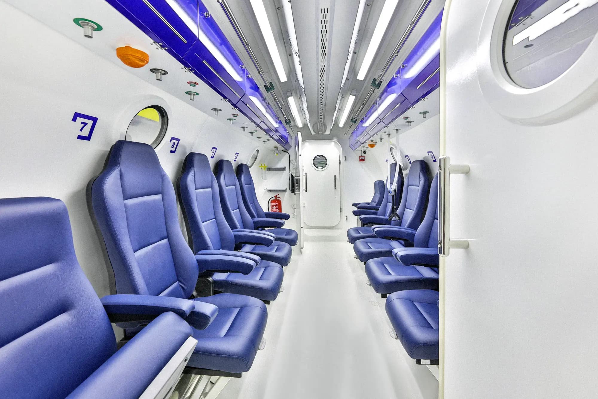 Interior aisle of a Hipertech multiplace chamber, numbered ergonomic seats down both sides and the inner lock door at the far end