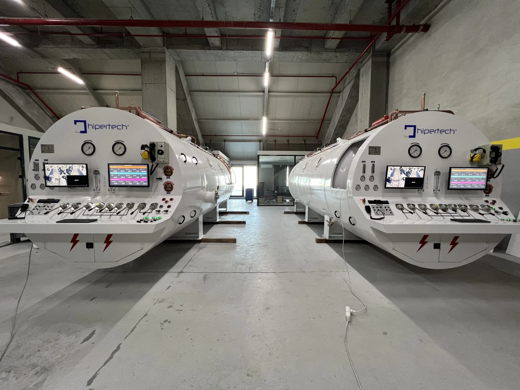 Two white Hipertech chambers standing side by side in a concrete building shell, control panels and gauges facing the camera, during installation