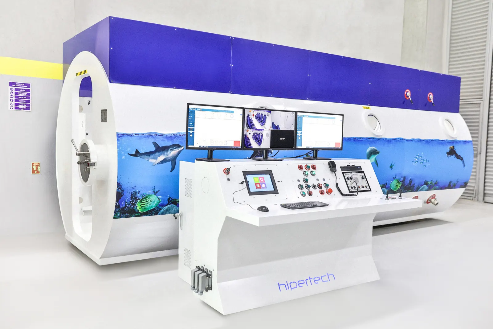 A Hipertech operator console with three screens standing in front of a Multiplace Omega finished in a blue undersea mural