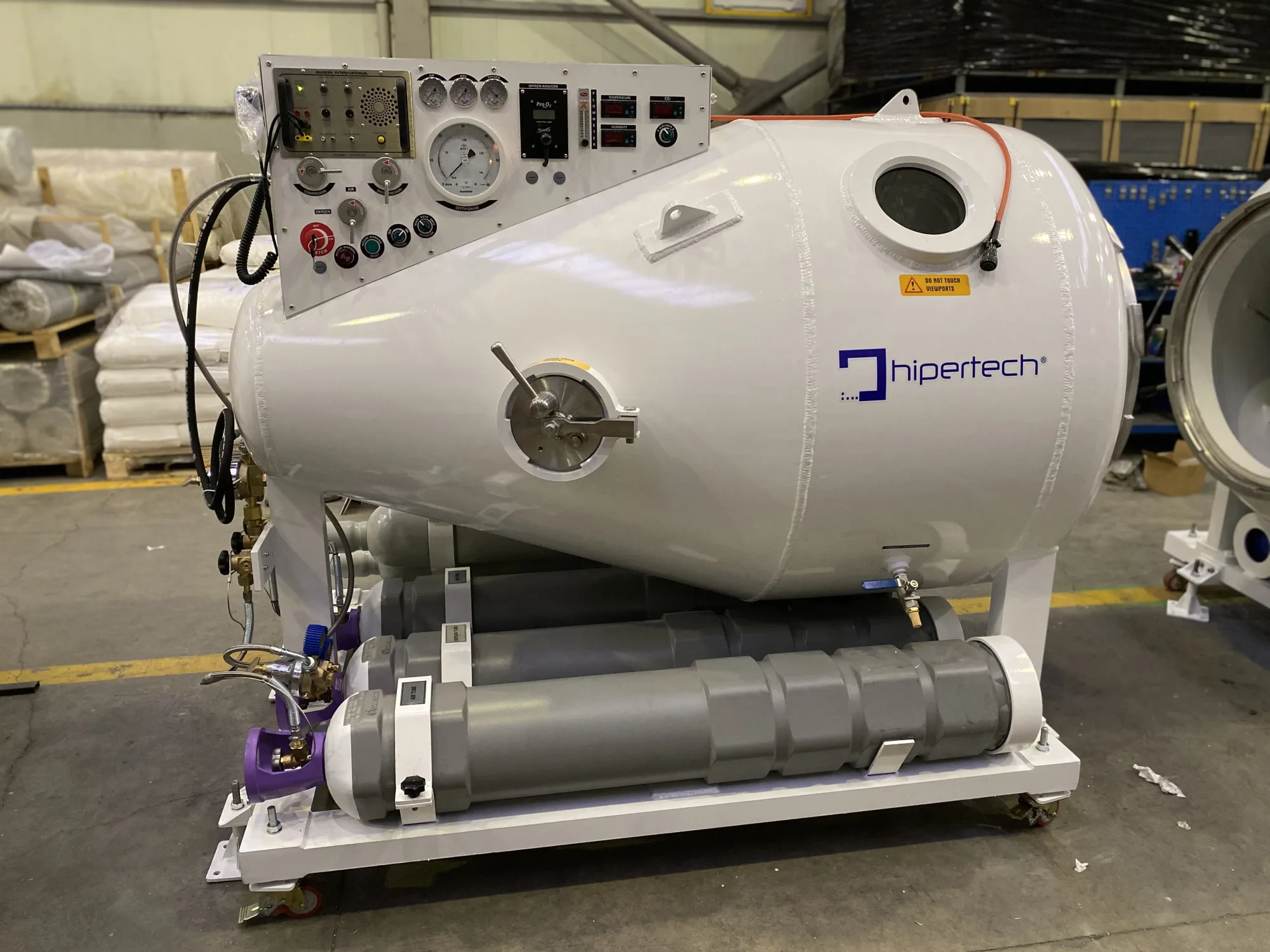 A Hipertech diving chamber on a white transport skid, composite gas cylinders slung under the frame and the operating console mounted on the angled face of the hull